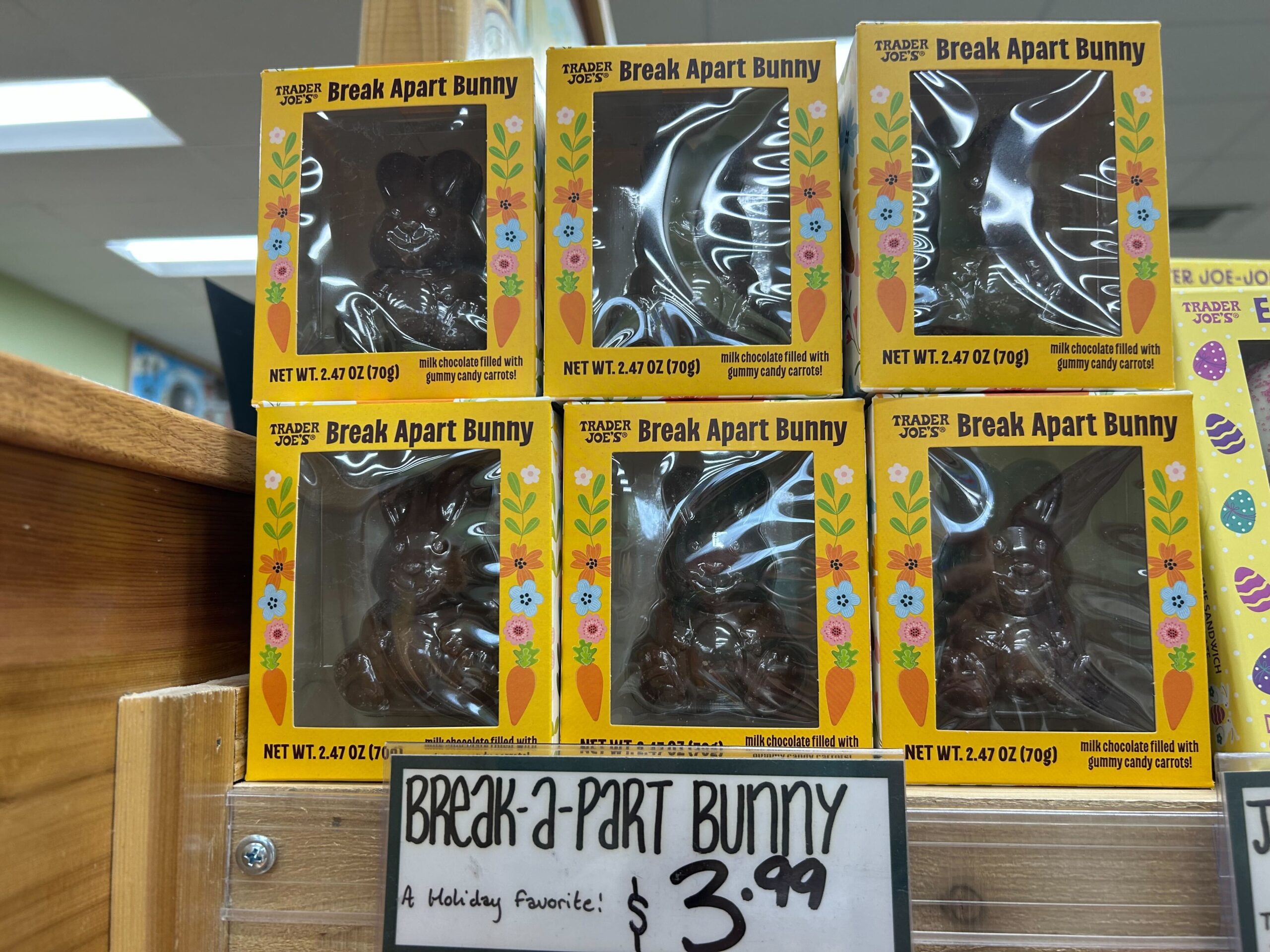 Boxes of Trader Joe’s Break Apart Bunny chocolates are displayed on a wooden shelf. A handwritten sign below reads “Break-a-part Bunny $3.99” with a note, “A Holiday Favorite.”