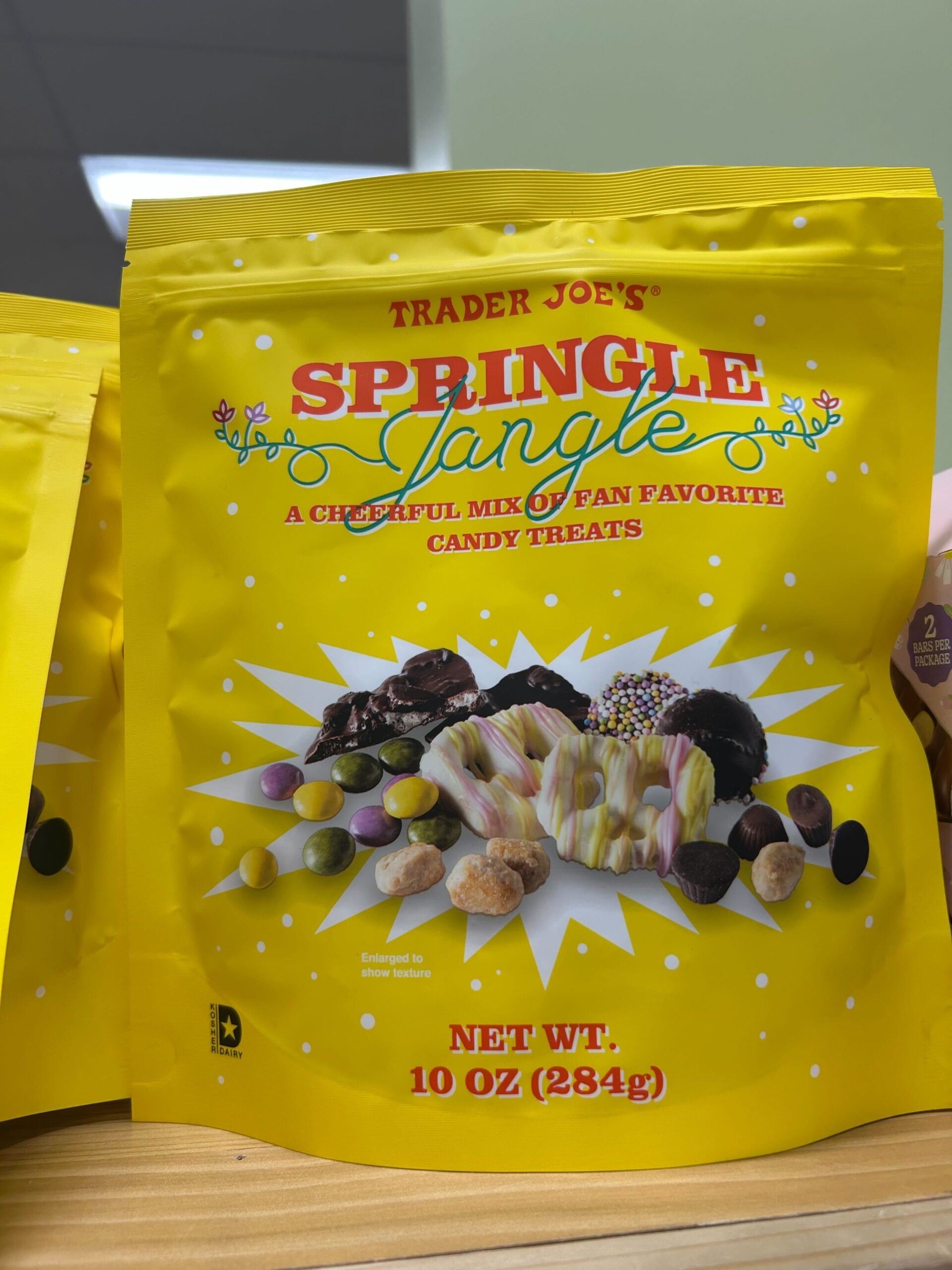 A yellow bag of Trader Joe’s Springle Jangle candy mix sits on a shelf. The bag features colorful candy illustrations and text that reads, "A cheerful mix of fan favorite candy treats," and has a net weight of 10 oz (284g).