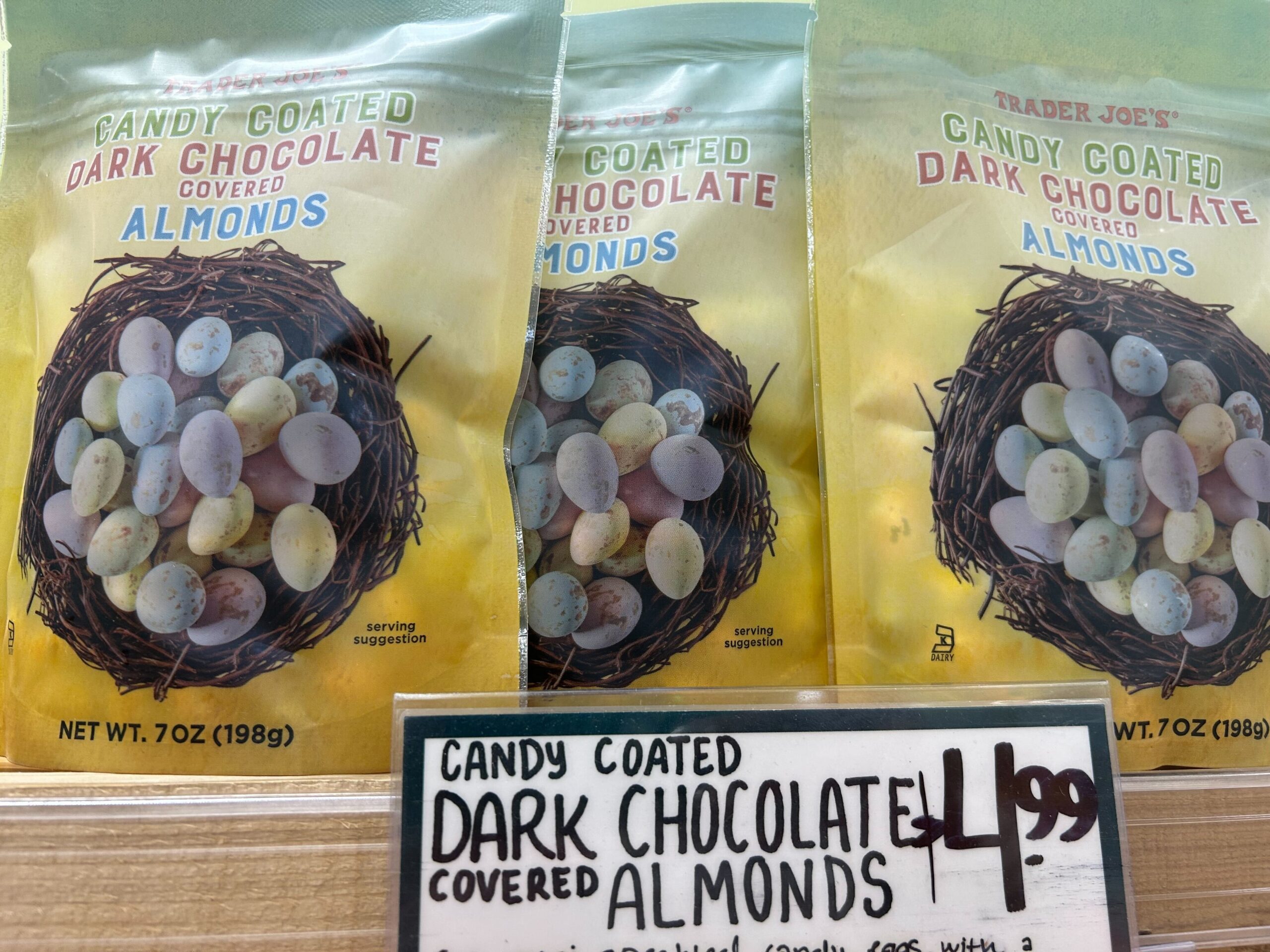 Bags of Trader Joe’s candy coated dark chocolate covered almonds, decorated like pastel speckled eggs in a nest, are displayed on a shelf with a price sign showing $4.99.