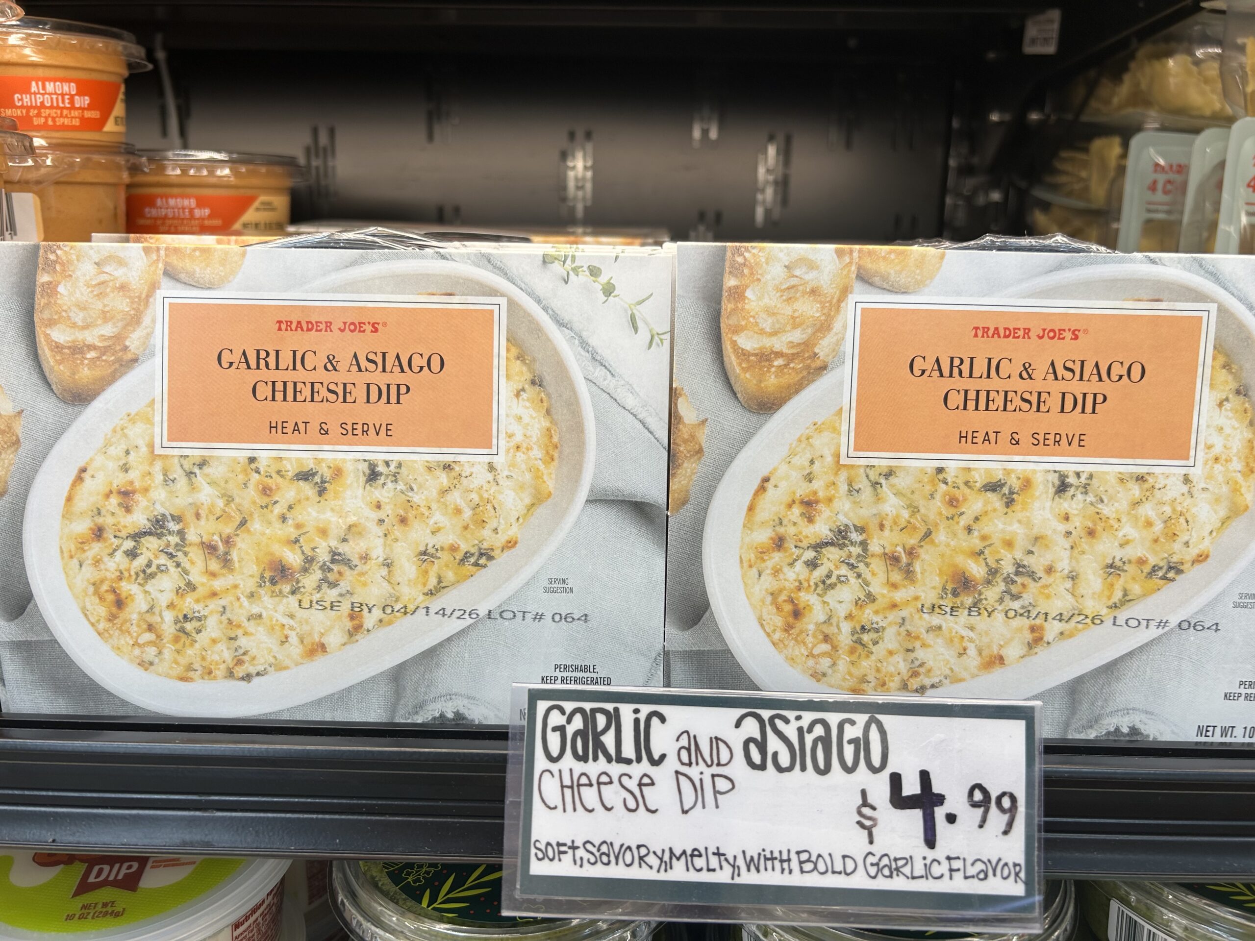 Two boxes of Trader Joe’s Garlic & Asiago Cheese Dip are displayed on a store shelf. A handwritten sign below reads: “Garlic and Asiago Cheese Dip $4.99. Soft, savory, melty, with bold garlic flavor.”