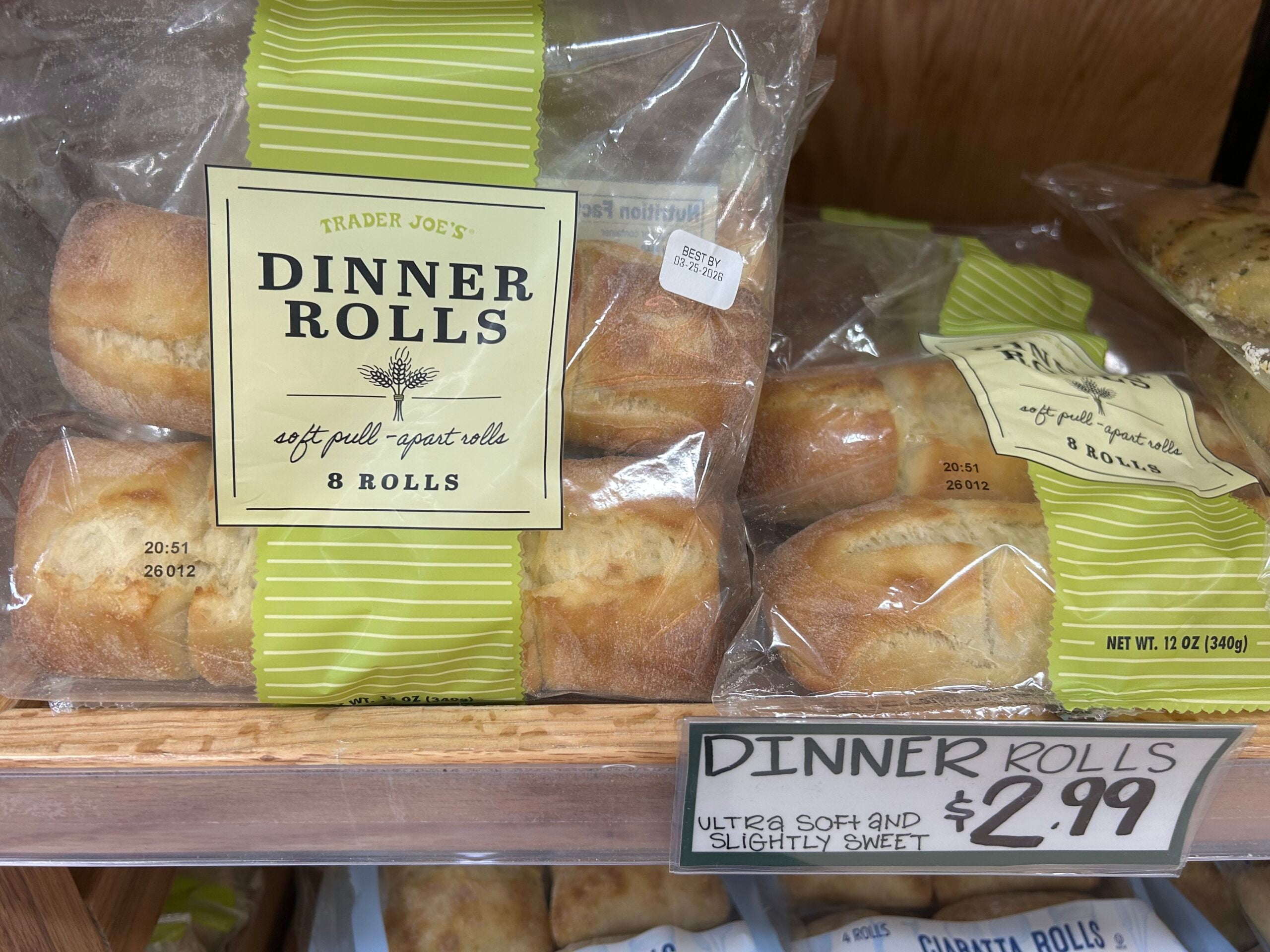 A package of Trader Joe’s Dinner Rolls labeled “soft pull-apart rolls, 8 rolls” sits on a shelf above a sign that reads: “Dinner Rolls $2.99, ultra soft and slightly sweet.”