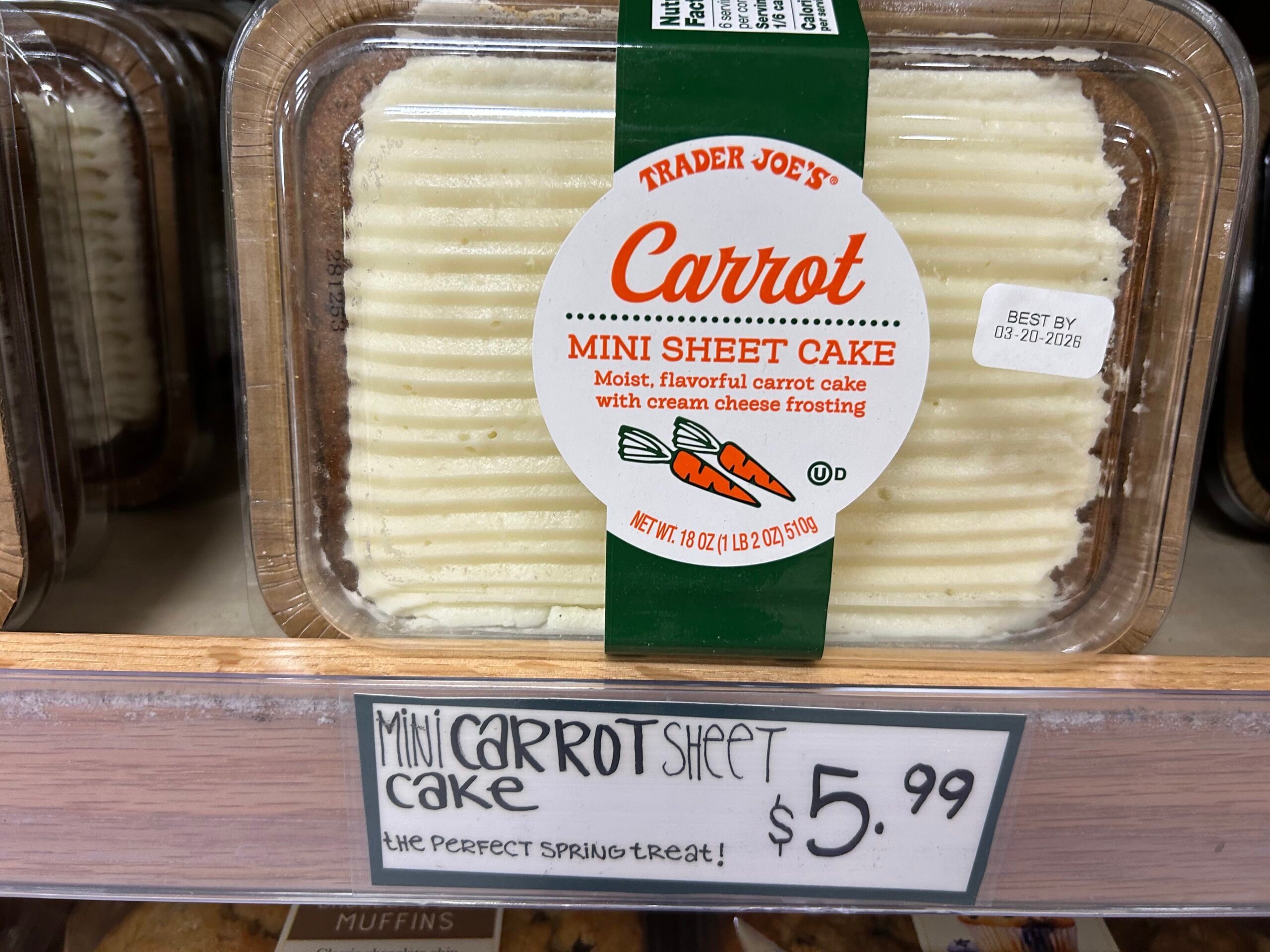 A package of Trader Joe’s Carrot Mini Sheet Cake with cream cheese frosting sits on a store shelf. A handwritten sign below reads "Mini Carrot Sheet Cake $5.99 the perfect spring treat!
