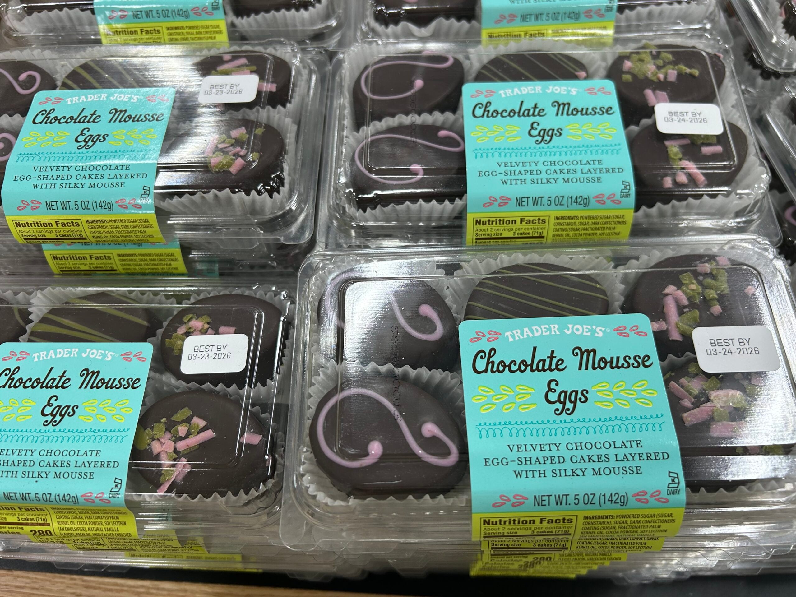 Plastic packages of Trader Joe’s Chocolate Mousse Eggs, each containing two chocolate egg-shaped desserts with pastel decorations, are stacked on display in a store. The yellow and blue label is visible on each package.