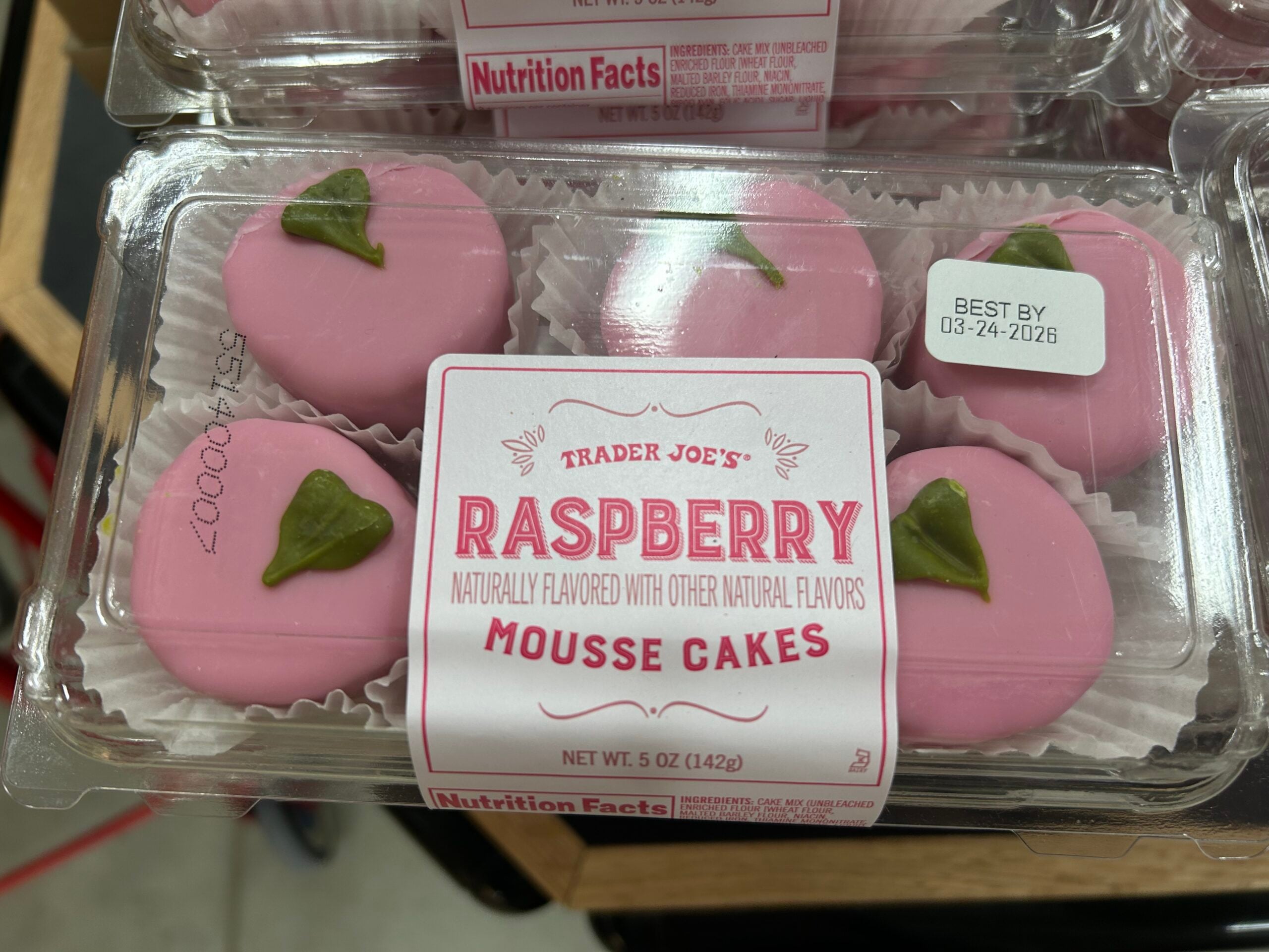 A clear plastic box of Trader Joe's Raspberry Mousse Cakes, pink and round with green leaf decorations on top, labeled "Best By 03-24-2026" and "Net Wt. 5 oz (142g)".