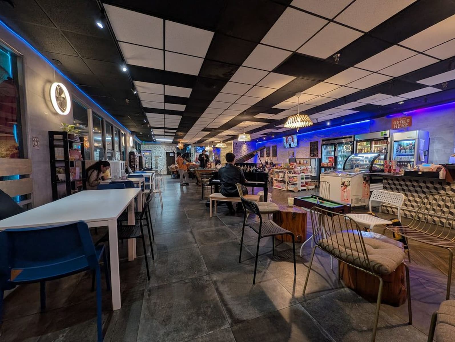 A modern café with a checkered ceiling, blue accent lighting, tables and chairs, a counter with desserts, and a few people sitting and ordering. The atmosphere is cozy and inviting.