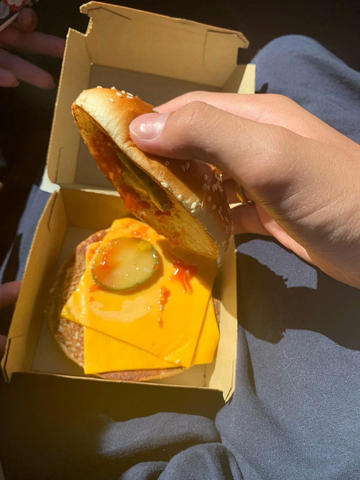 A hand lifts the top bun of a cheeseburger in a cardboard box, revealing a slice of cheese, two pickles, and ketchup on the patty. Sunlight highlights the burger and the person’s lap under the box.