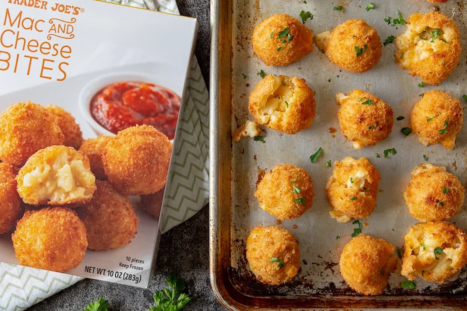 A baking tray with golden brown mac and cheese bites is next to a Trader Joe’s box labeled “Mac and Cheese Bites.” Some bites are garnished with herbs, and the box shows a serving suggestion with marinara sauce.
