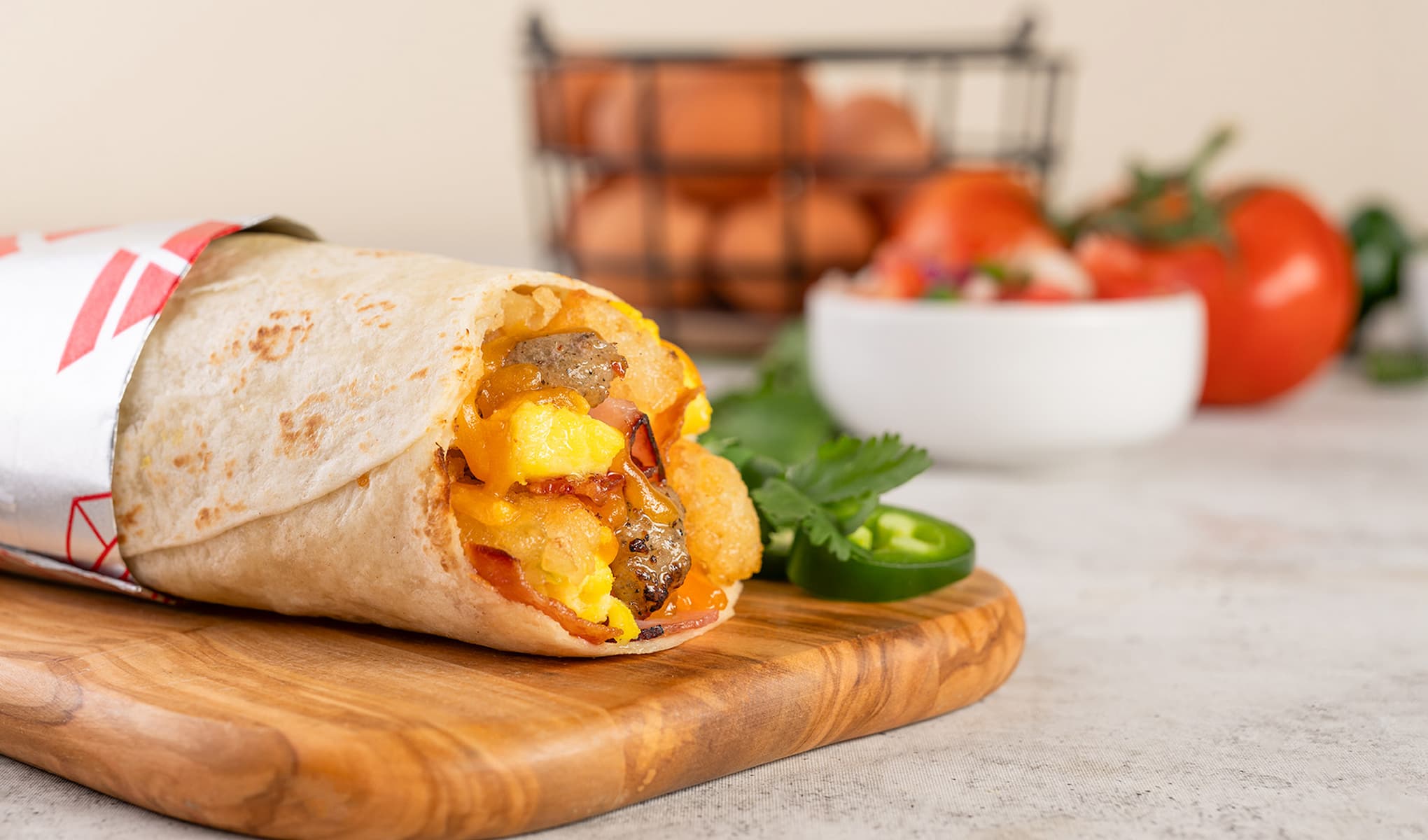 A breakfast burrito filled with eggs, potatoes, cheese, and sausage on a wooden board, with sliced jalapeño and cilantro. In the background, eggs in a wire basket and tomatoes near a bowl of salsa.