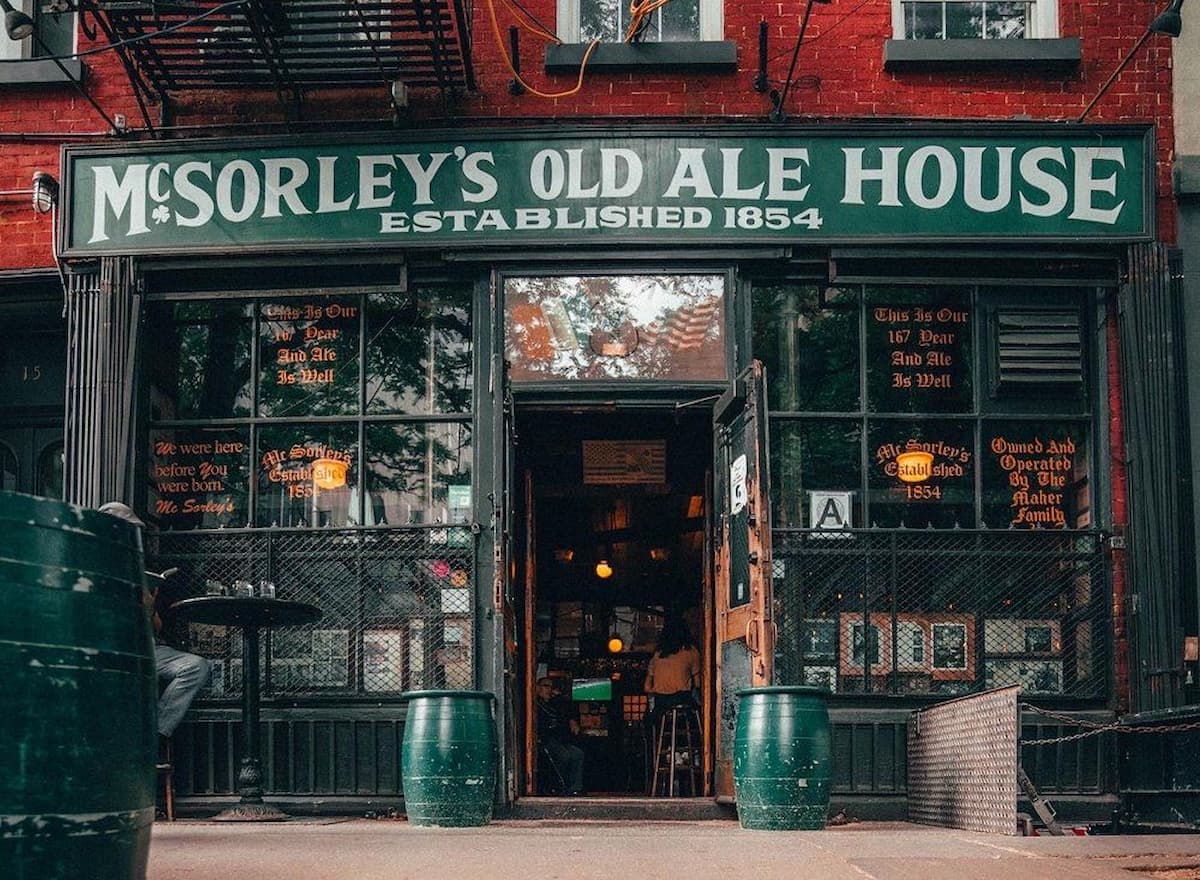 The entrance of McSorley’s Old Ale House, a historic pub established in 1854, with green barrels outside, large front windows, and a partially open door revealing a dimly lit interior with patrons.