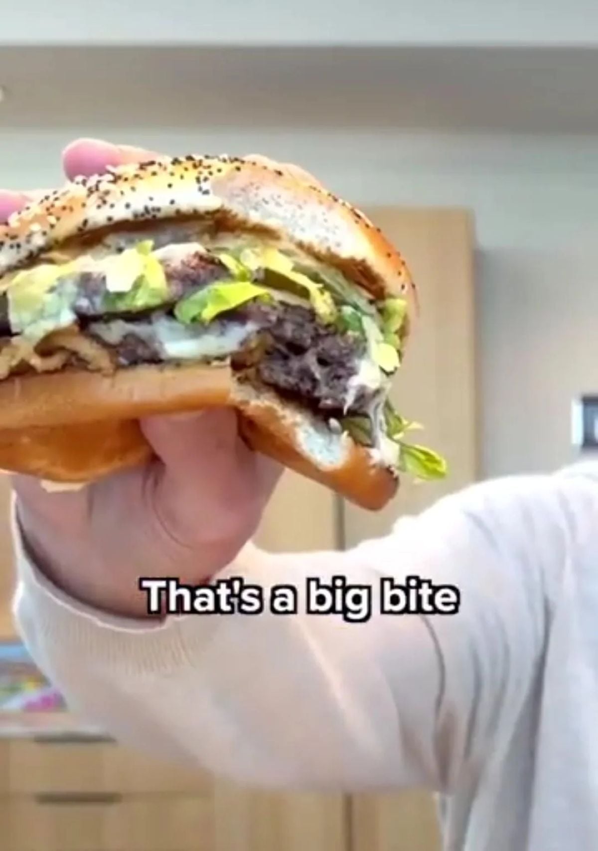 A person holds up a large, partially eaten hamburger with lettuce, cheese, onions, and sauce. The text on the image reads, "That's a big bite.