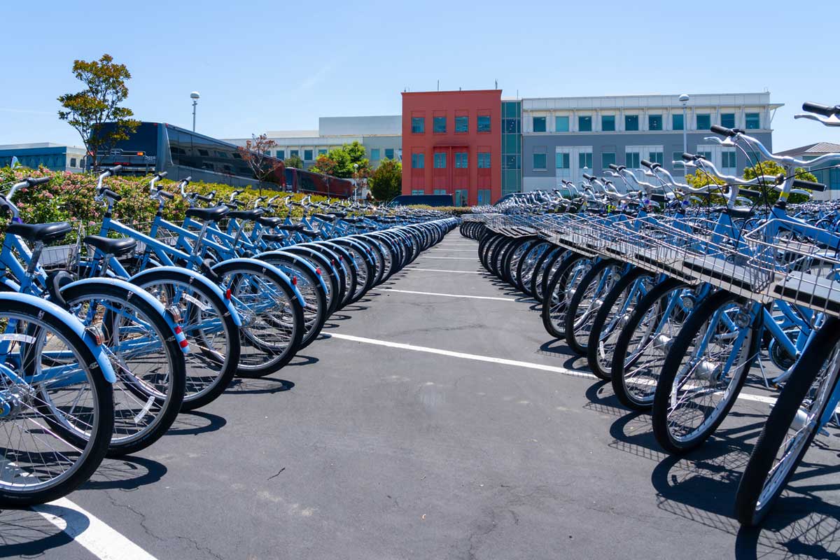 Rows of blue bicycles are lined up in a parking lot on a sunny day, with a red and blue building in the background, showing quiet streets after the 2026 layoffs impacted local businesses.