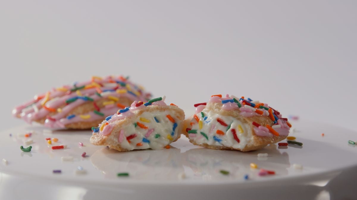 A close-up of a cream-filled cookie cut in half, topped with pink icing and colorful sprinkles, sits on a white surface with more sprinkles scattered around.