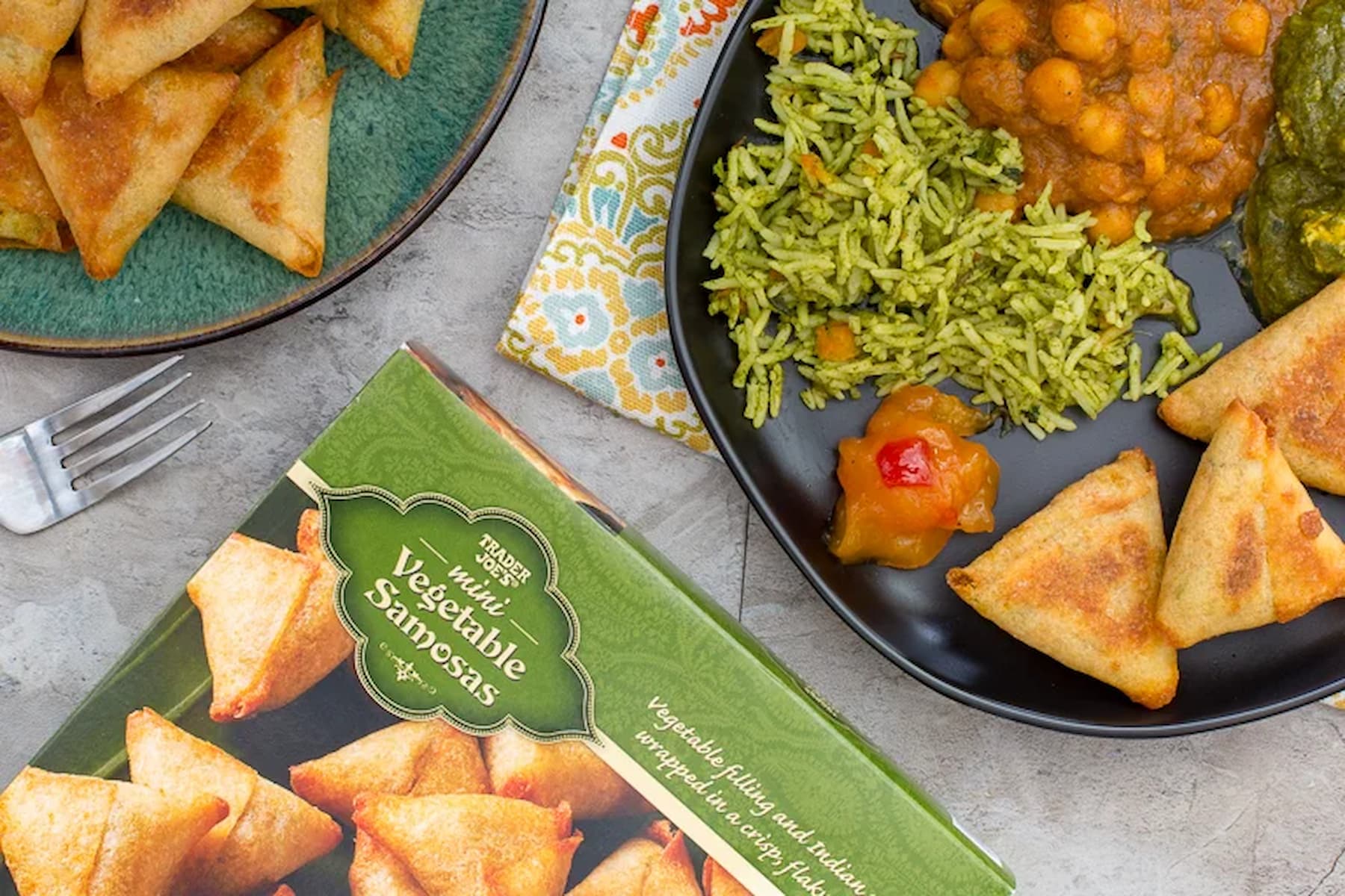 A plate with green rice, chickpea curry, spinach, vegetable samosas, and chutney sits beside a box labeled "Vegetable Samosas" and a plate of more samosas; a fork and napkin are nearby.