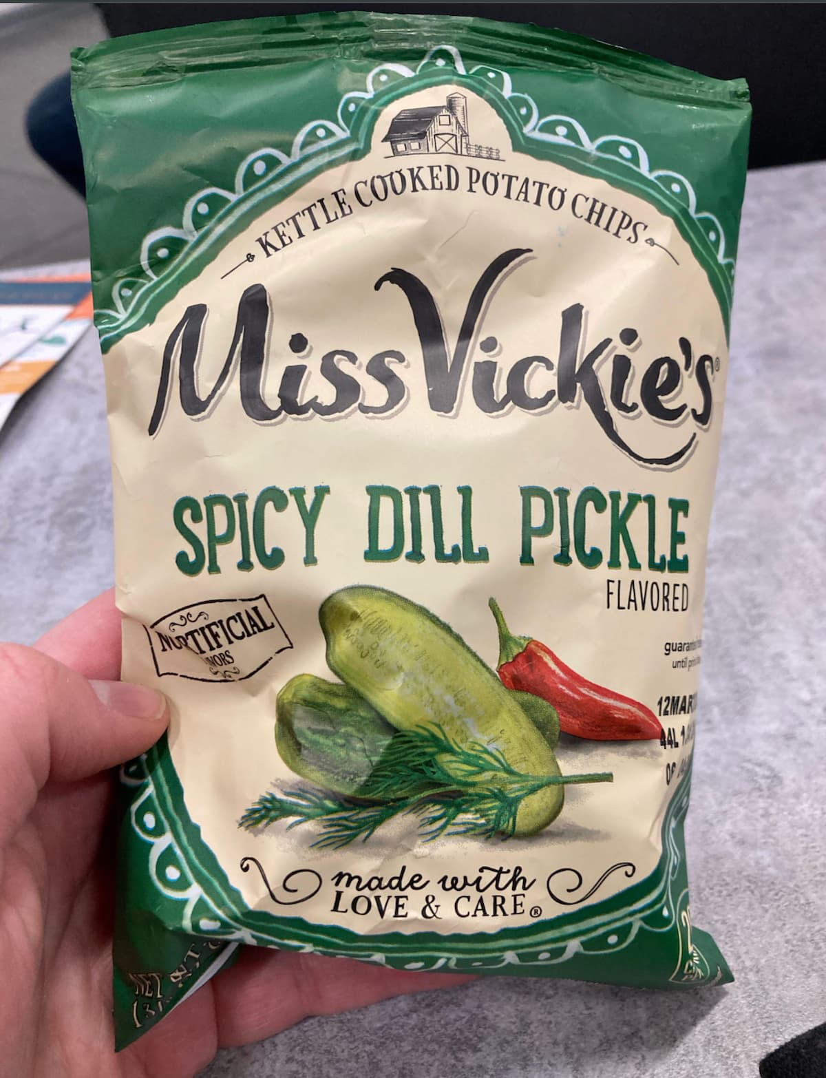 A hand holds a bag of Miss Vickie's Spicy Dill Pickle flavored kettle cooked potato chips. The bag is green and cream-colored, featuring images of pickles, dill, and a red chili pepper.