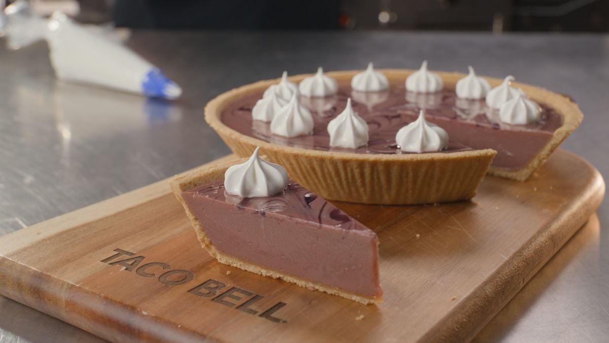 A chocolate cream pie with a graham cracker crust is on a wooden board labeled "Taco Bell." A slice is cut and placed in front, and both the pie and slice are topped with dollops of whipped cream.