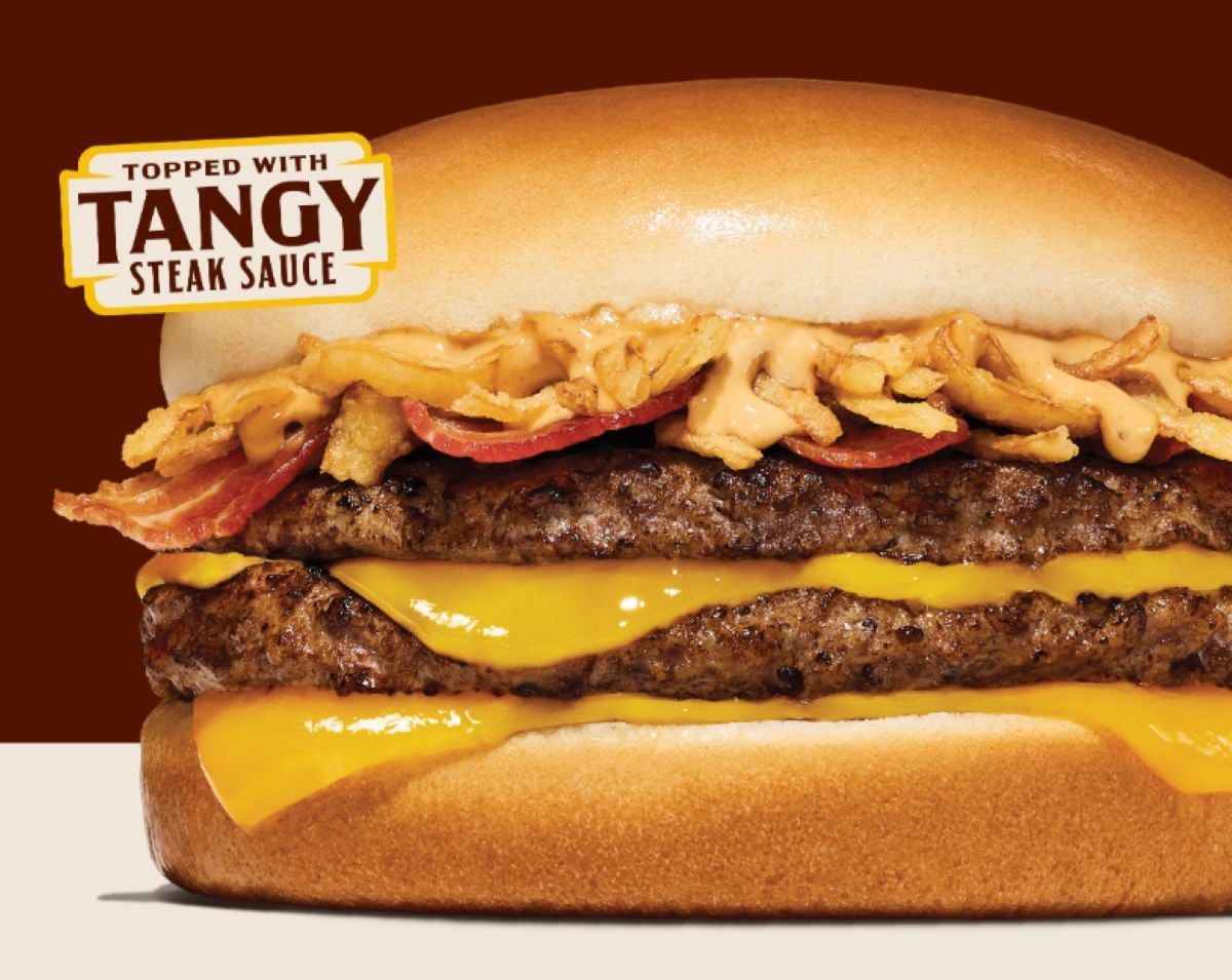 A close-up of a cheeseburger from a new fast-food menu, stacked with two beef patties, melted cheddar cheese, crispy onions, bacon, and tangy steak sauce on a soft bun. A label in the corner reads "Topped with Tangy Steak Sauce.