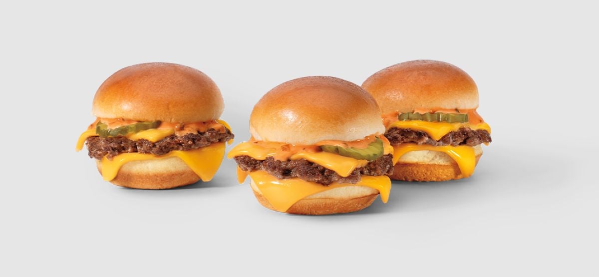 Three cheeseburgers from the new fast-food menu feature beef patties, melted cheddar cheese, pickles, and sauce in shiny toasted buns, all arranged on a light gray background.