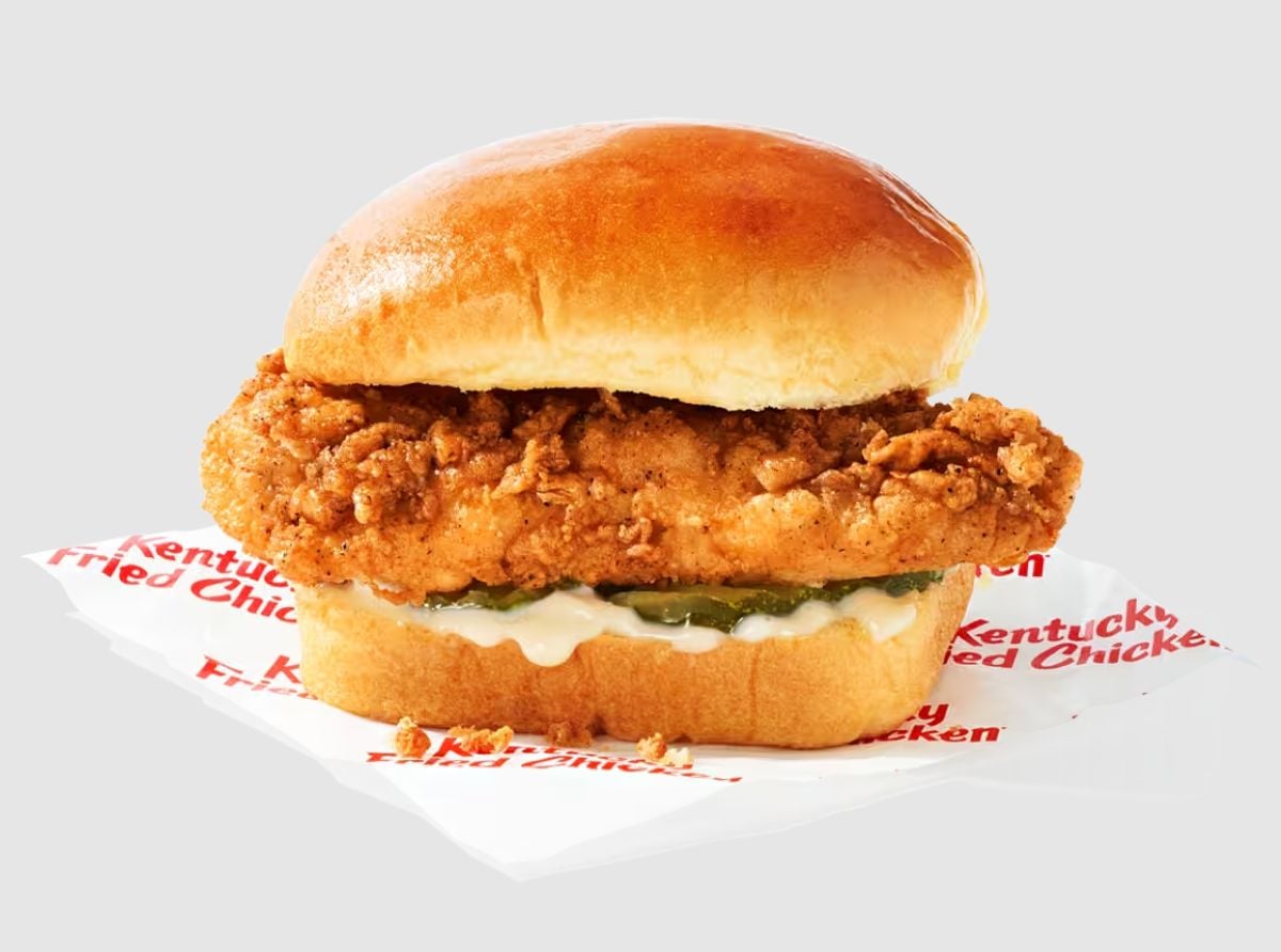 A crispy fried chicken fillet sandwich with pickles and mayonnaise in a shiny brioche bun, featured on the new fast-food menu, placed on a KFC-branded wrapper against a light gray background.