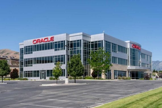 A modern, three-story office building with "Oracle" signage on its facade and large glass windows stands over a mostly empty parking lot&mdash;perhaps hinting at the impact of the 2026 layoffs. Trees and mountains rise in the background under a clear blue sky.