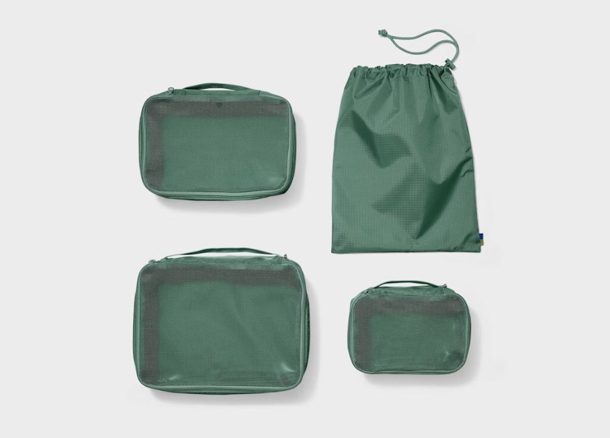 Four green travel packing cubes and a drawstring bag are arranged on a white background—perfect for spring break. The cubes come in various sizes with mesh tops for visibility, while the solid bag features a cinch closure.