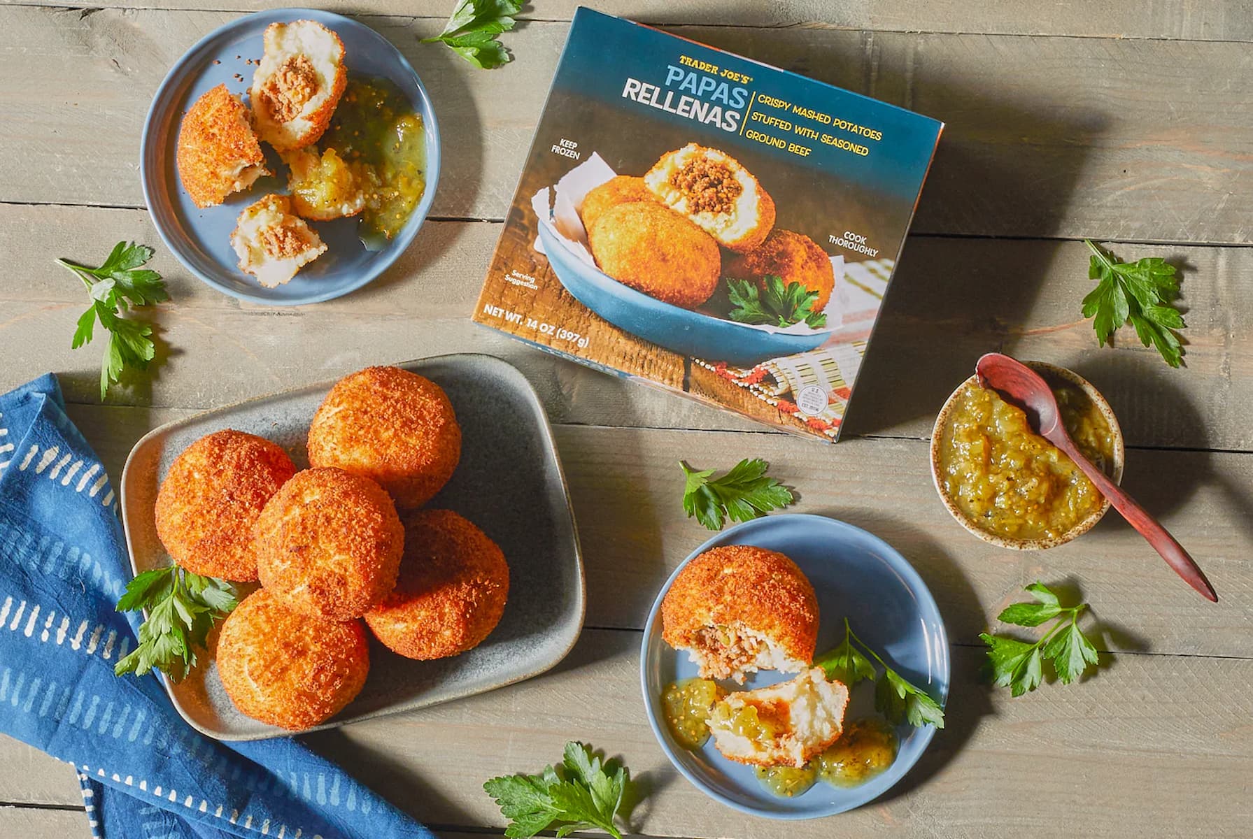 Five breaded potato balls, some cut open, are served on small plates with green sauce. A box labeled “Papas Rellenas” and a bowl of sauce with a spoon are nearby on a wooden table, garnished with parsley.