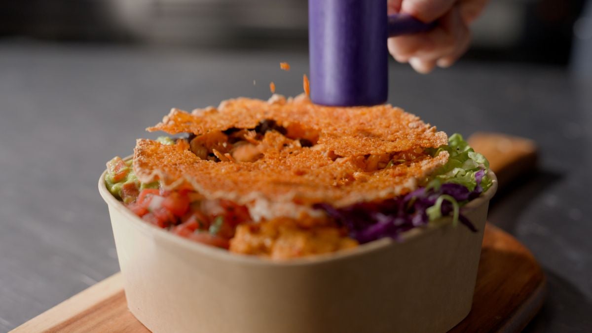 A close-up of a bowl filled with colorful vegetables and toppings, with someone sprinkling orange seasoning from a small purple container over the crispy top layer.