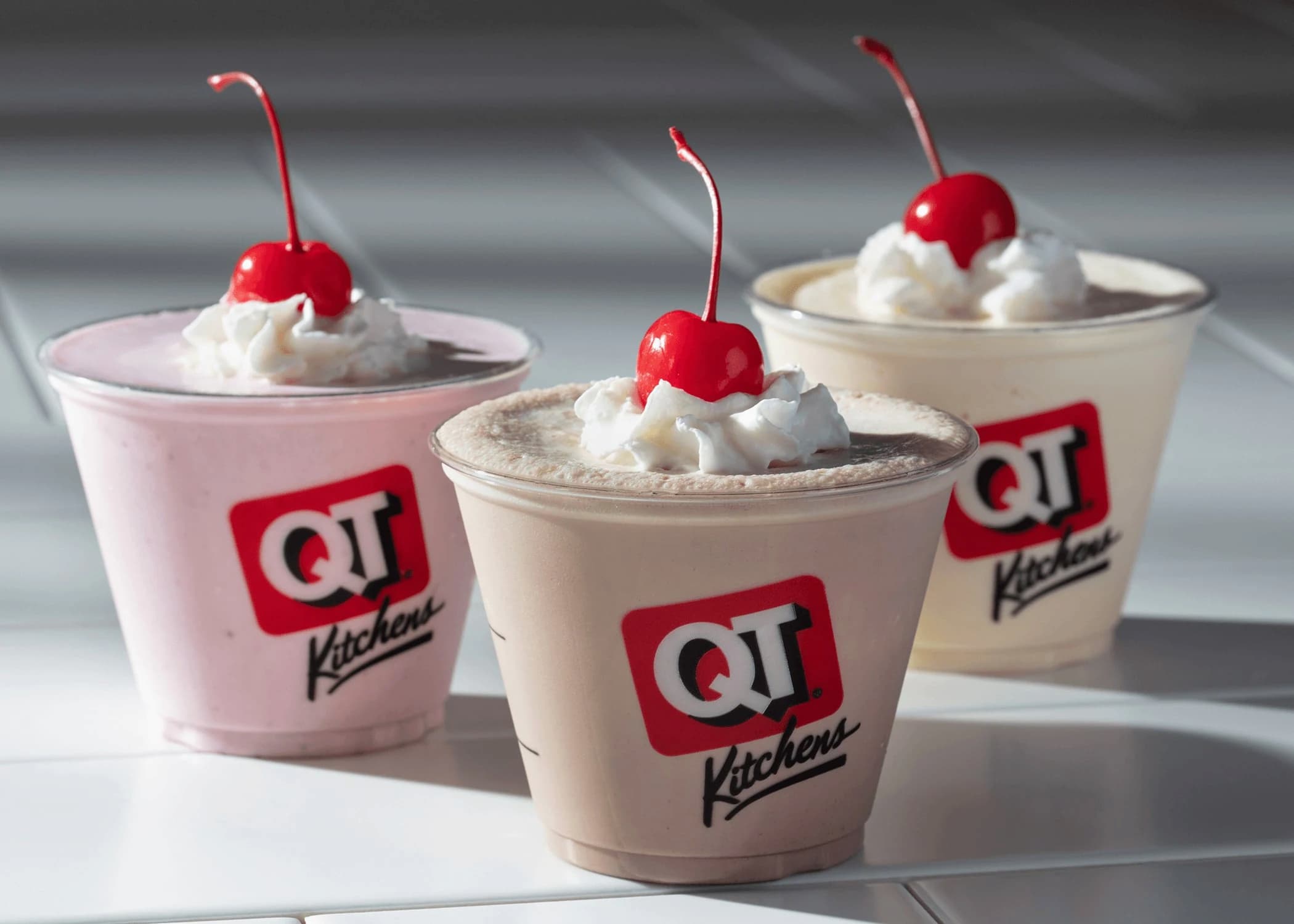 Three QT Kitchens milkshakes in clear cups topped with whipped cream and cherries, with chocolate, vanilla, and strawberry flavors, placed on a white tiled surface.