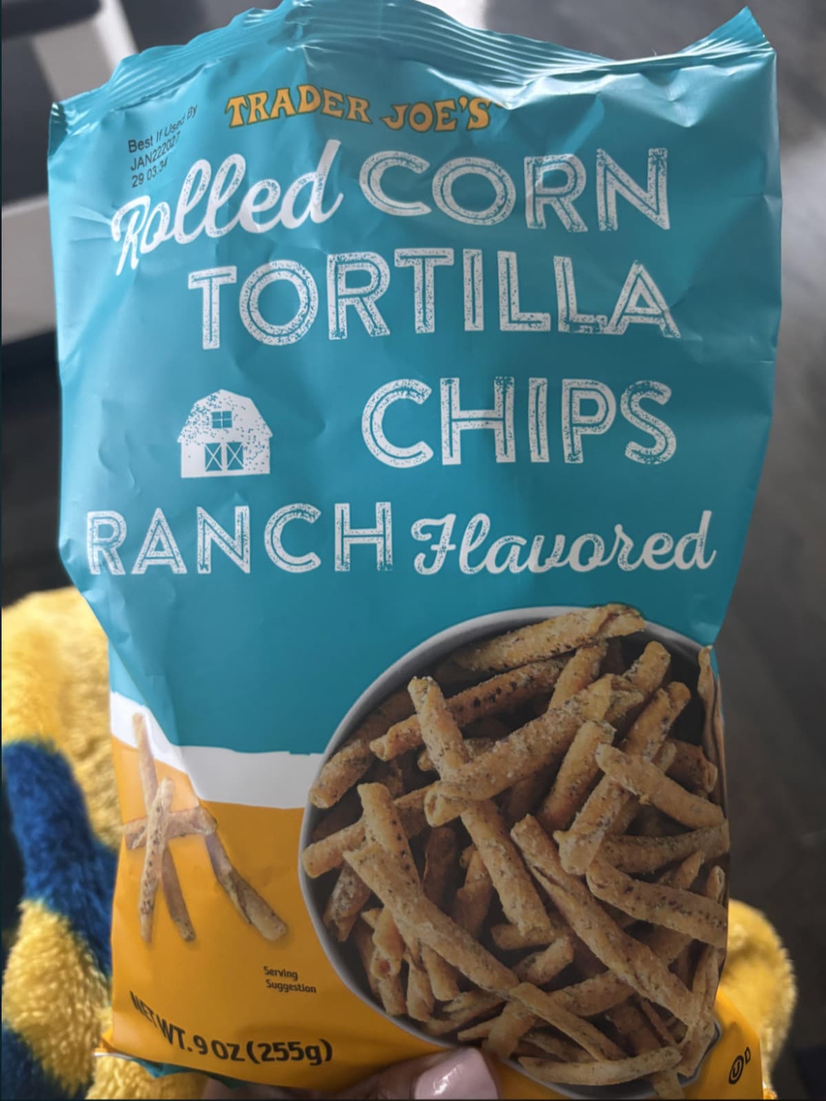 A hand holds a blue bag of Trader Joe’s Rolled Corn Tortilla Chips, Ranch Flavored. The bag features an image of the chips in a bowl and displays the net weight as 9 oz (255g).