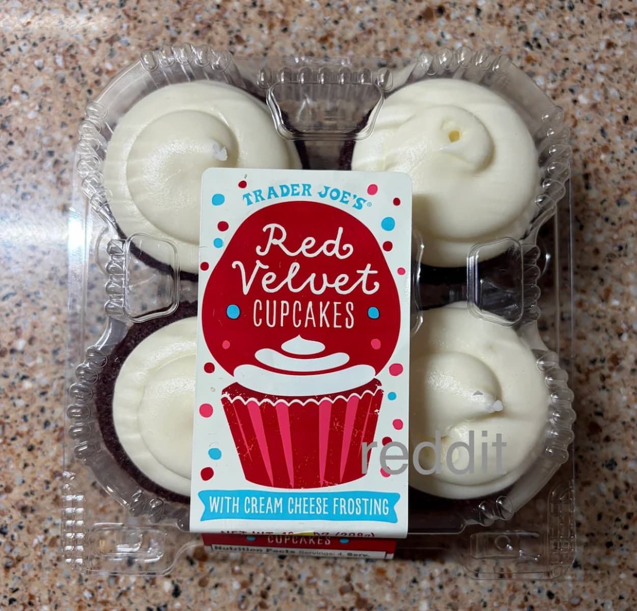 A clear plastic container holds four red velvet cupcakes with white cream cheese frosting. The label reads "Trader Joe’s Red Velvet Cupcakes with Cream Cheese Frosting" and features a red cupcake illustration.
