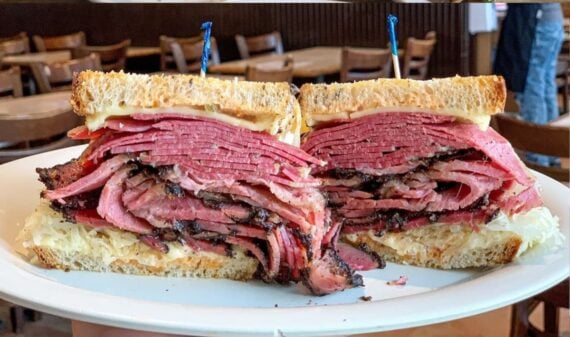 A plate holds a large deli sandwich, sliced in half, stacked high with layers of pastrami and dark crusted meat on rye bread, with sauerkraut and creamy dressing. Wooden picks hold each half together.