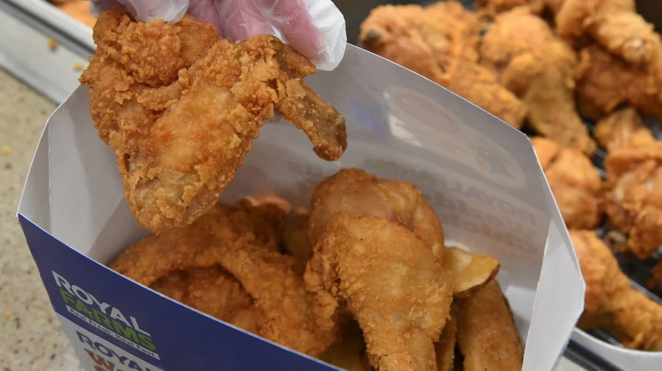 A gloved hand holds a piece of crispy fried chicken above a box filled with several pieces of fried chicken. The box is blue and white with visible text on the side.