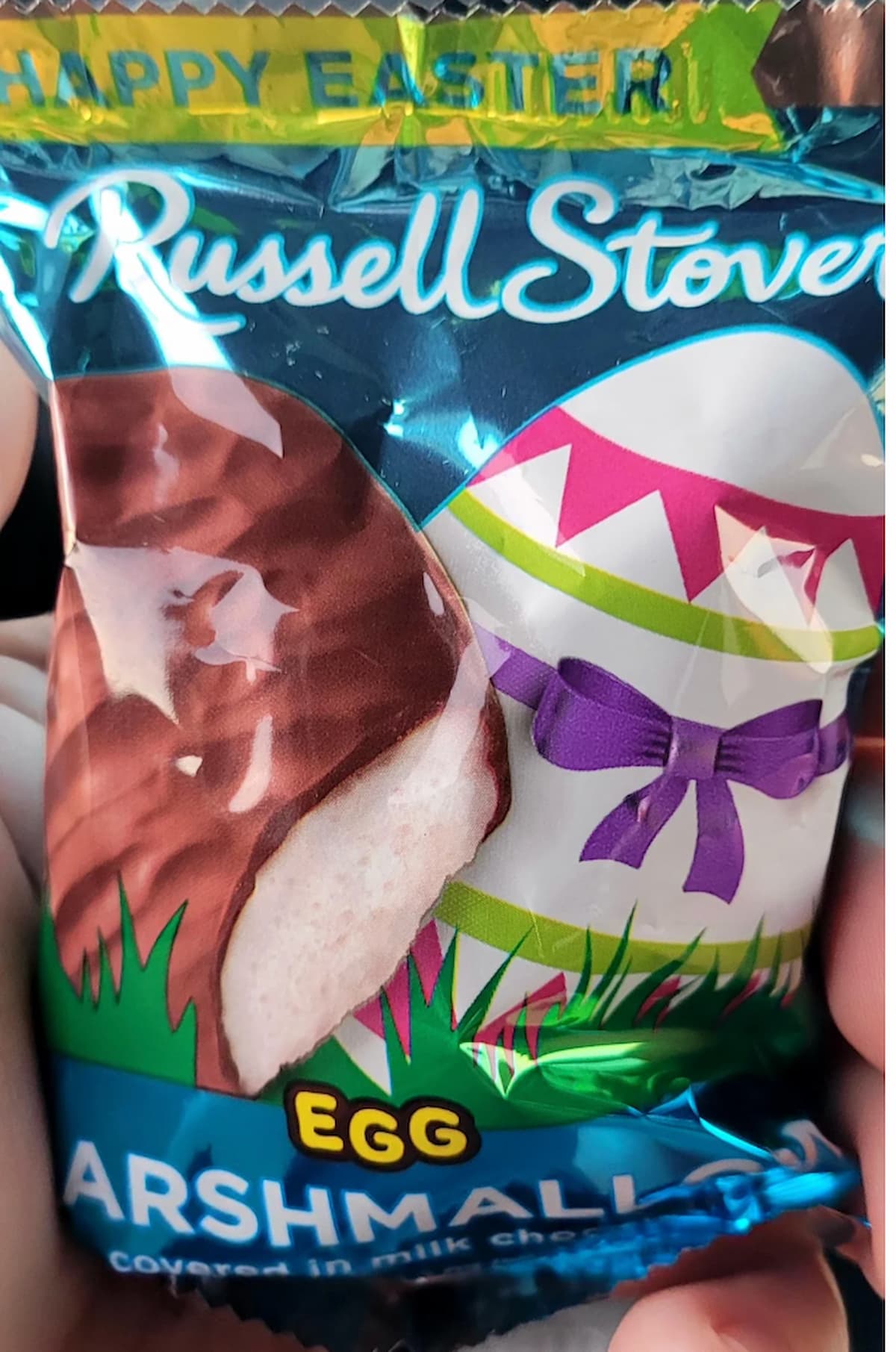 A close-up of a Russell Stover marshmallow egg candy in a blue wrapper, featuring an image of a chocolate-covered marshmallow egg and a decorated Easter egg with a purple bow.