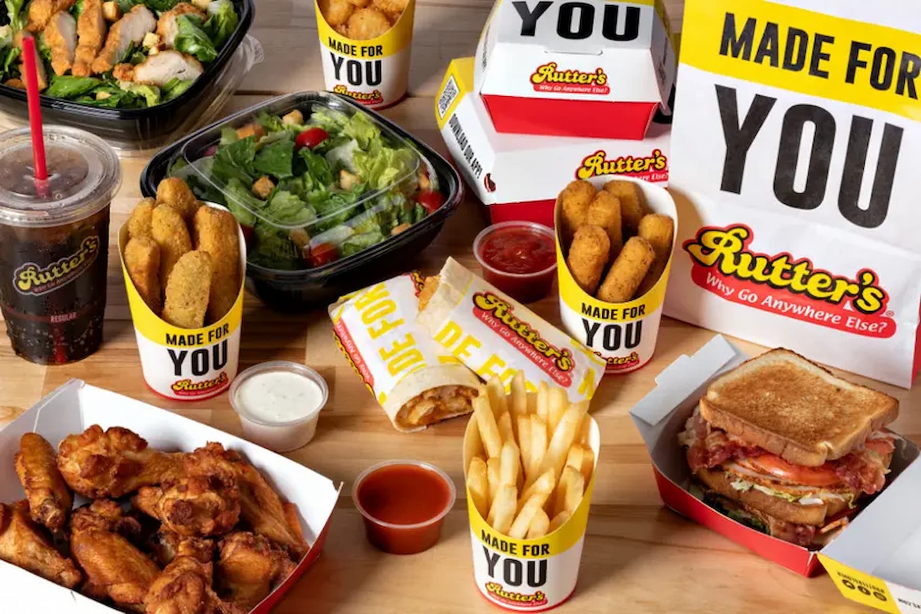 A variety of fast food items including chicken wings, mozzarella sticks, French fries, salads, a sandwich, dipping sauces, a fountain drink, and packaging with the Rutter’s logo on a wooden table.