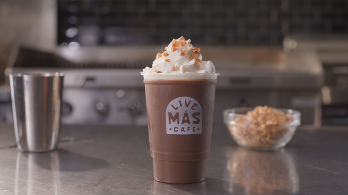 A chocolate frozen drink topped with whipped cream and caramel drizzle sits on a metal counter. The cup reads "LIVE MÁS CAFÉ." In the background are a metal cup and a bowl of cereal.