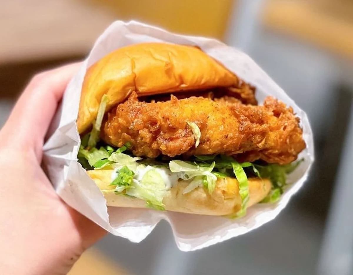 A hand holds a fried chicken sandwich with crispy chicken, shredded lettuce, and a soft bun, wrapped partially in white paper—perfect for enjoying while checking out the latest March Madness restaurant deals.