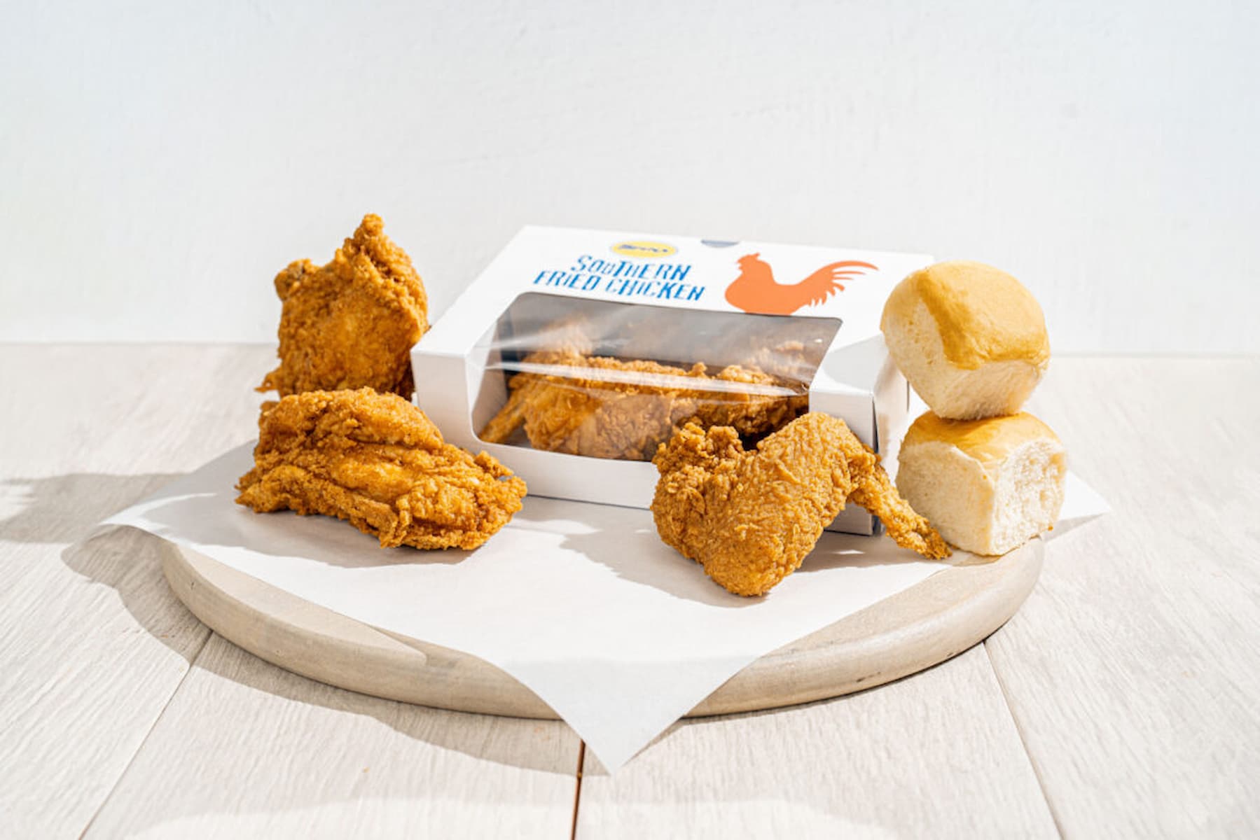 A serving of Southern fried chicken pieces, two bread rolls, and a box with more fried chicken pieces, arranged on a round wooden board with a white paper liner.