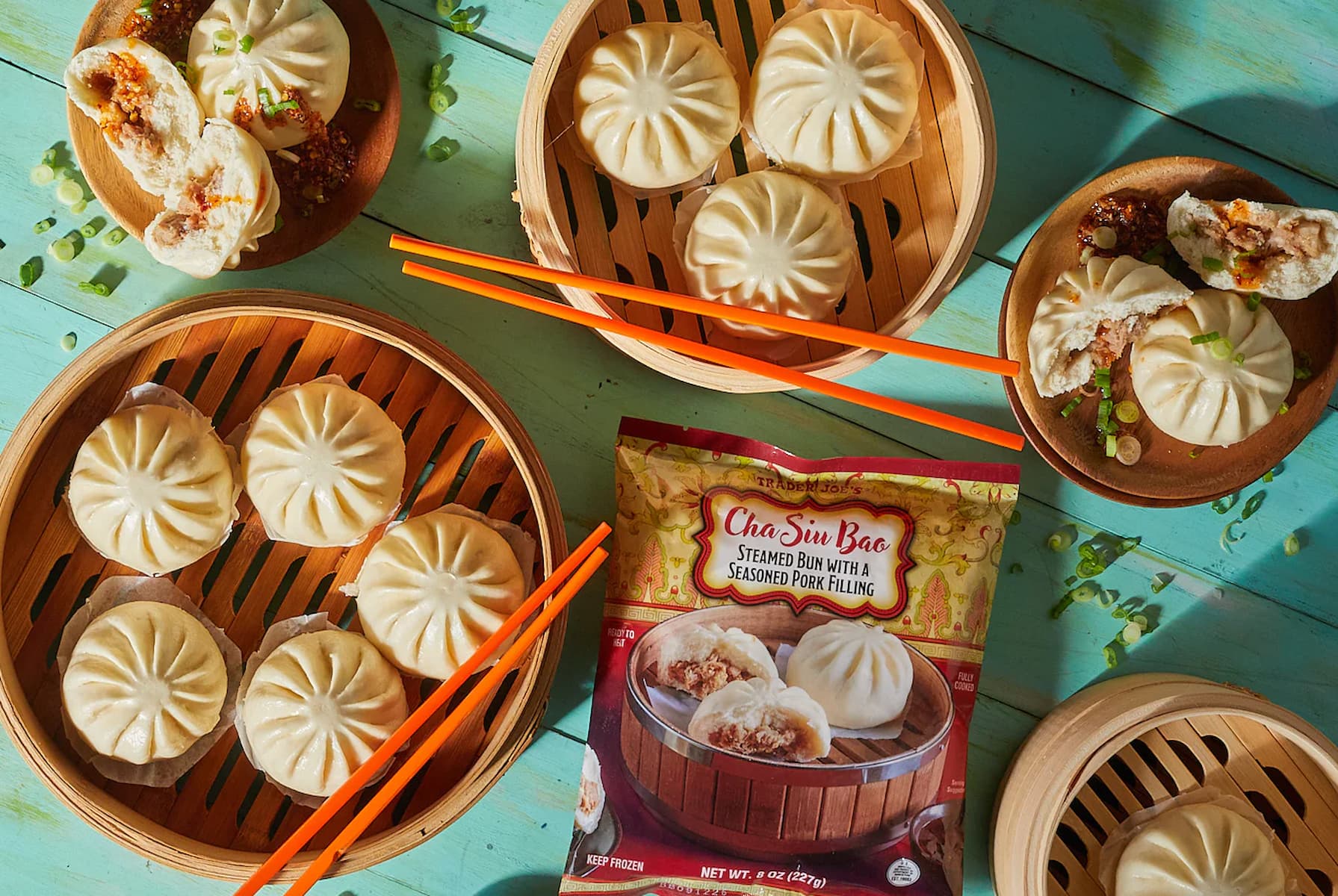 Top-down view of bamboo steamers filled with steamed pork buns, some buns cut open, garnished with green onions. A packet labeled "Cha Siu Bao Steamed Bun with Seasoned Pork Filling" is also visible. Orange chopsticks included.