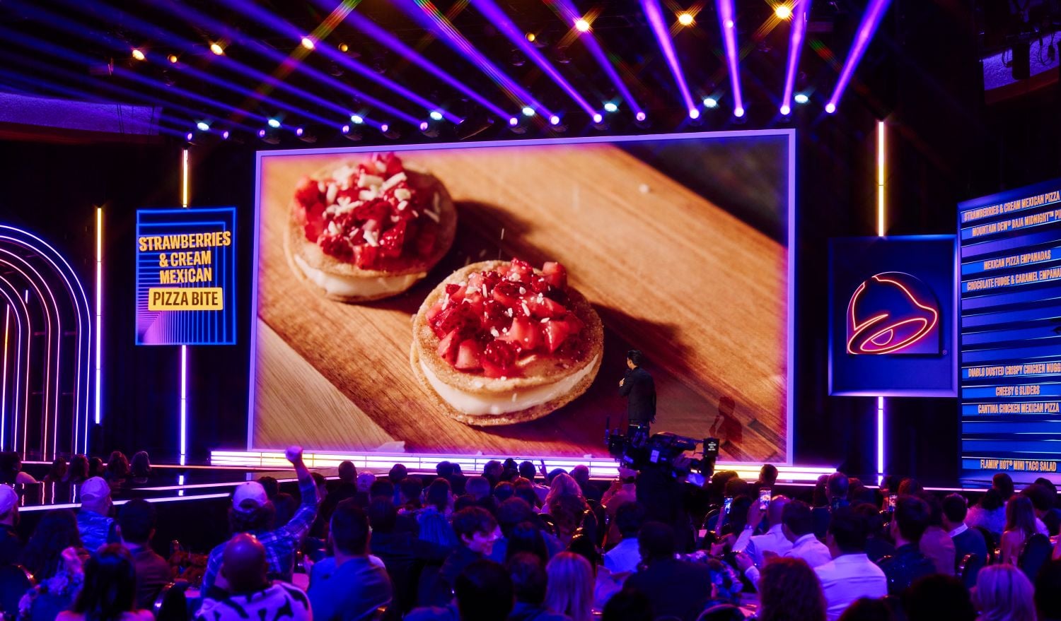 A large audience watches a stage presentation showing a big screen with an image of two strawberry and cream Mexican pizza bites, while colorful lights illuminate the venue.
