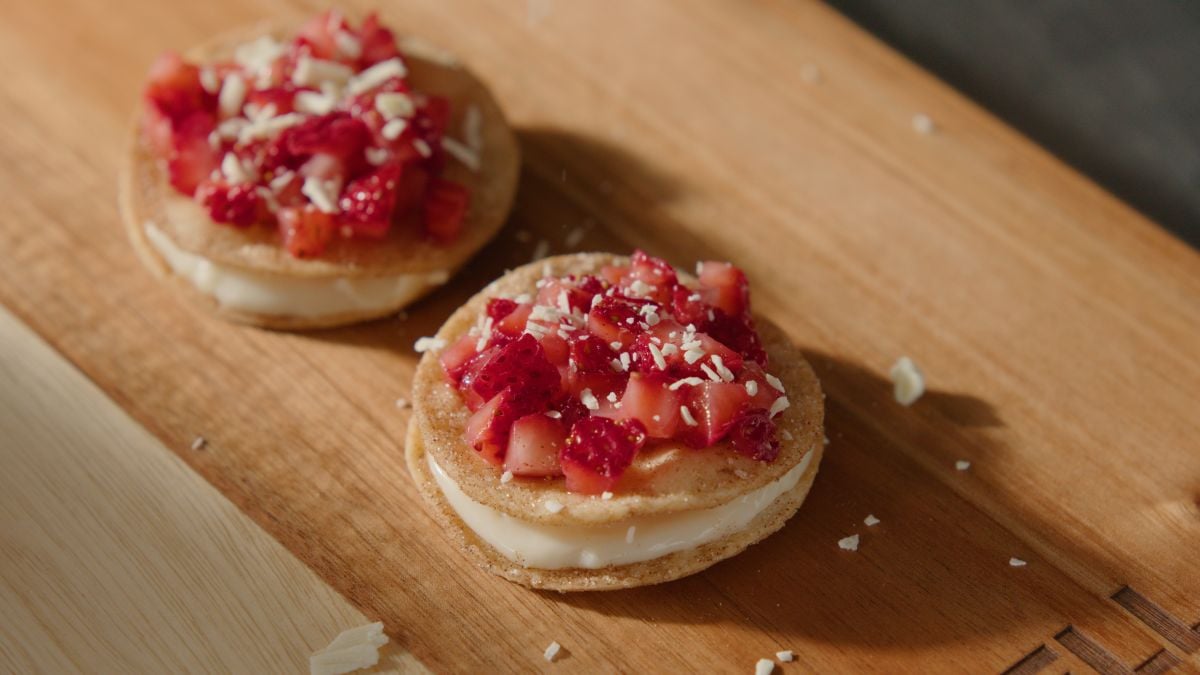 Two round cookie sandwiches with a layer of cream filling are topped with diced strawberries and sprinkled with white chocolate shavings, placed on a wooden board.