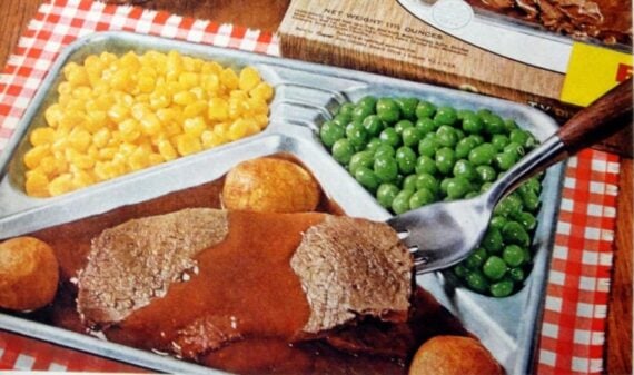 A vintage TV dinner with sections of corn, green peas, two potato puffs, and sliced roast beef with brown gravy, served on a metal tray atop a red and white checkered tablecloth.