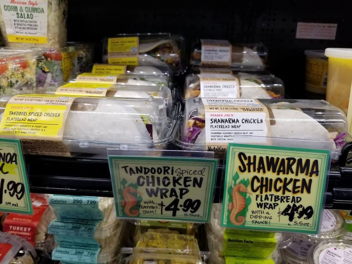 Plastic containers of tandoori spiced chicken wraps and shawarma chicken flatbread wraps are displayed on a grocery store shelf with handwritten price tags, reading $4.99.