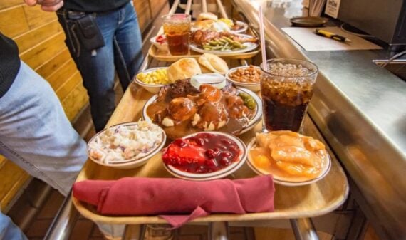 A wooden tray filled with plates of comfort food, including meat with gravy, biscuits, mashed potatoes, corn, beans, fruit desserts, coleslaw, and a glass of soda, is being carried by a person in jeans.