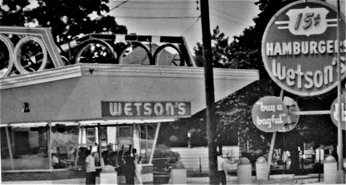 Black-and-white photo of a retro hamburger stand called "Wetson's," with a large sign advertising 15¢ hamburgers and the slogan "Buy 'em by the bagful." Several people stand near the entrance.