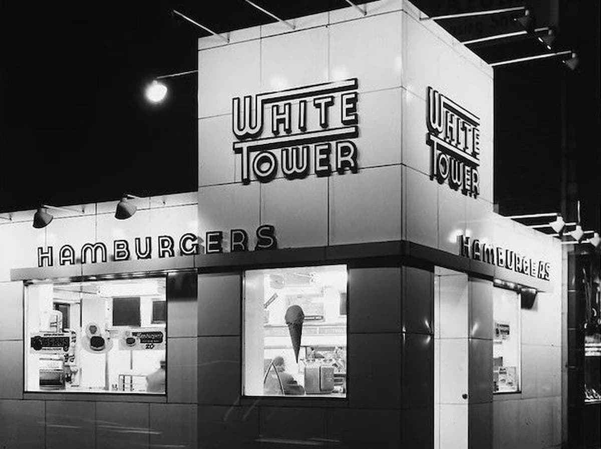 A vintage, black-and-white photo of a White Tower restaurant at night, with illuminated signs reading "White Tower" and "Hamburgers." The building has a shiny, white tiled exterior and large windows showing the interior.