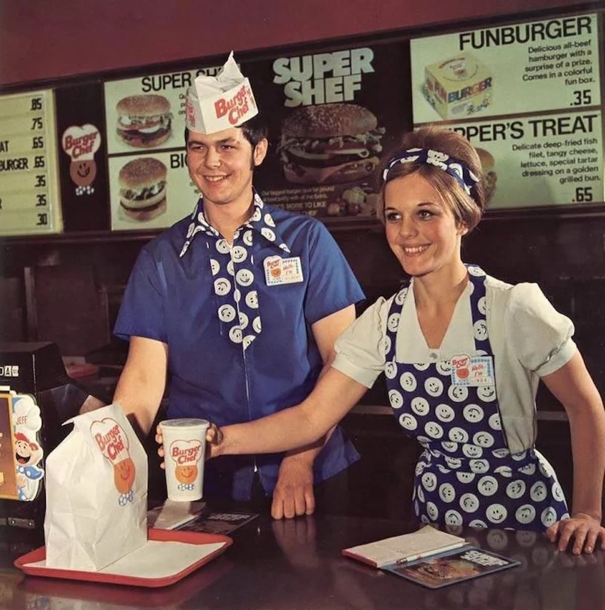 Two smiling fast-food employees in blue uniforms with smiley face patterns serve food and drinks at a Burger Chef counter, with menu boards displaying burger options in the background.