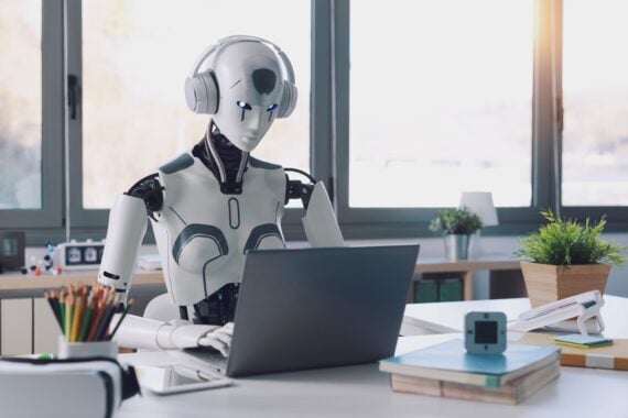 A humanoid robot wearing headphones sits at a desk, typing on a laptop. The desk has books, a pencil holder, and a potted plant. Large windows in the background let in natural light.
