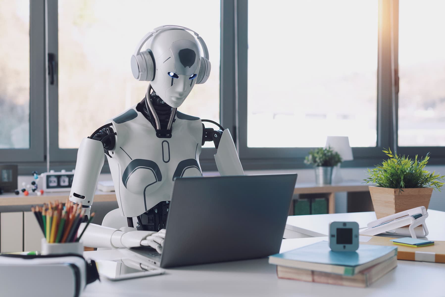 A humanoid robot wearing headphones sits at a desk, typing on a laptop. The desk has books, a pencil holder, and a potted plant. Large windows in the background let in natural light.