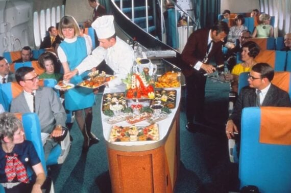 Flight attendants and a chef serve gourmet food and drinks to passengers seated in a spacious, retro airplane cabin with blue seats. Passengers appear well-dressed, chatting and enjoying the luxurious service.