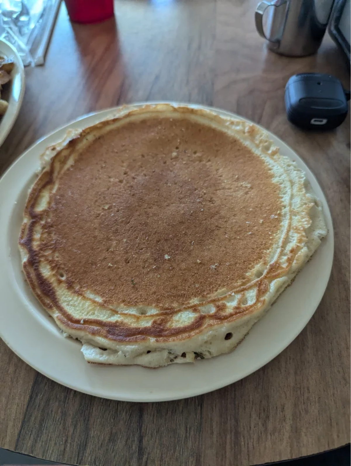 A large, golden-brown pancake sits on a white plate on a wooden table. Nearby are a red plastic cup, a metal creamer, and a black car key fob.