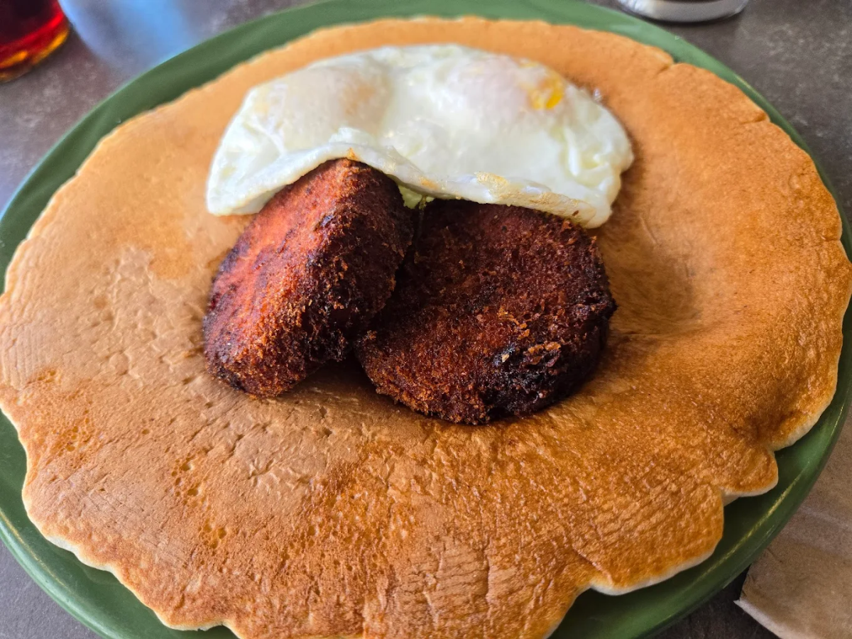 A large pancake on a green plate topped with two browned sausage patties and a sunny-side-up egg.